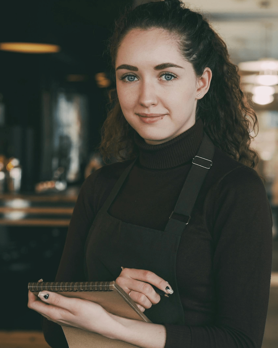 Portrait of a smiling young woman in a black turtleneck and apron, holding a brown notebook in a cafe setting.